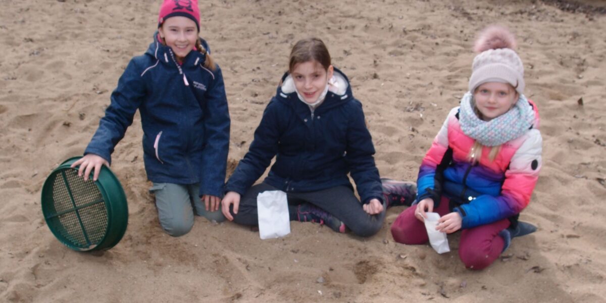 Drei Kinder in Jacken und Hüten sitzen im Sand. Eines hält ein grünes Sieb, zwei halten kleine weiße Säckchen. Im Hintergrund sind große Felsen zu sehen.