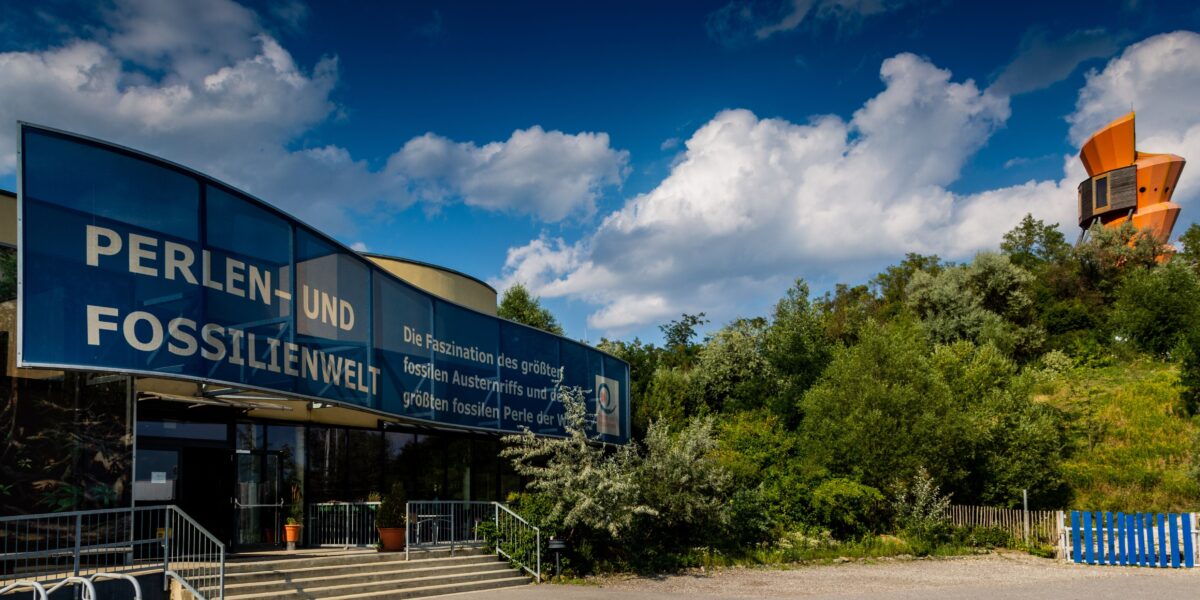 Ein Gebäude mit der Aufschrift "Perlen- und Fossilienwelt" steht neben einer Grünanlage unter einem blauen Himmel mit Wolken; im Hintergrund ist ein orangefarbener Aussichtsturm zu sehen.