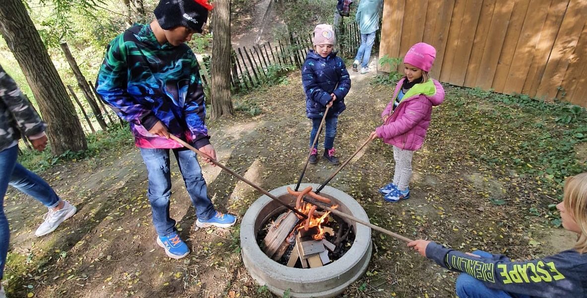 Vier Kinder stehen in einem Waldstück in der Nähe eines Holzzauns und eines Gebäudes um eine kleine Feuerstelle im Freien und halten lange Stöcke über die Flammen.