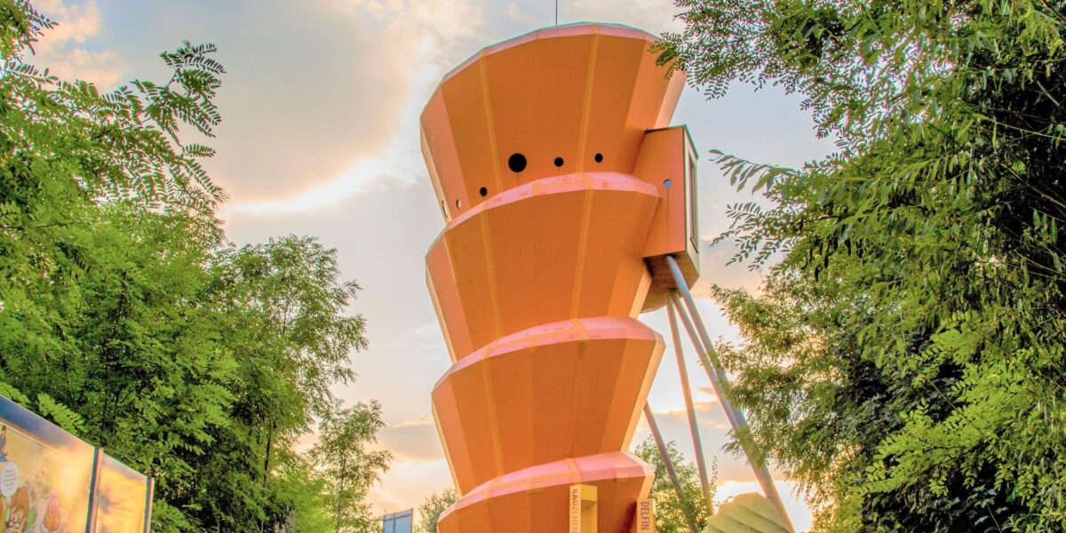 Ein orangefarbener, spiralförmiger Aussichtsturm steht zwischen grünen Bäumen unter einem teilweise bewölkten Himmel.