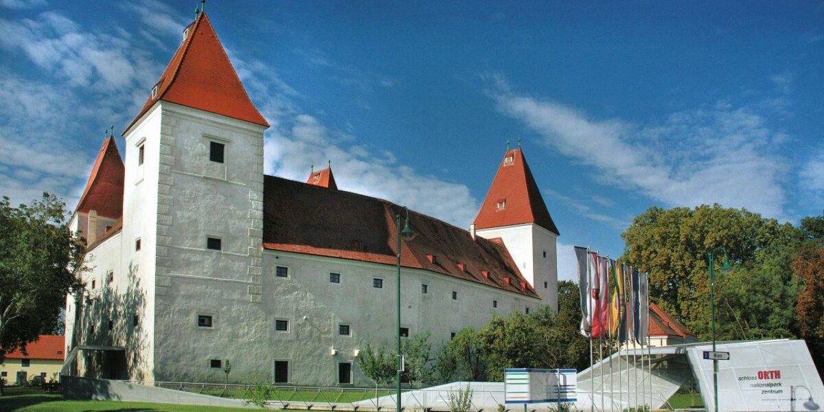 Ein weißes mittelalterliches Schloss mit roten Ziegeldächern und Ecktürmen, mit einem modernen Glaseingang, vor dem an einem sonnigen Tag mehrere Fahnen wehen.