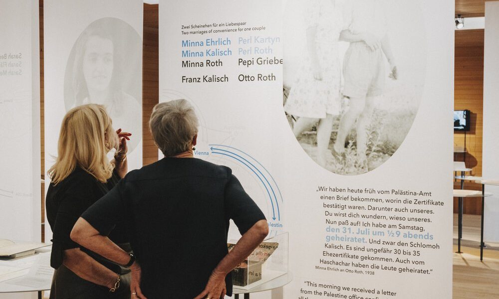 Zwei Frauen stehen vor einer Ausstellungstafel mit historischem Text, Namen und einem Schwarz-Weiß-Foto eines Paares.