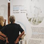 Zwei Frauen stehen vor einer Ausstellungstafel mit historischem Text, Namen und einem Schwarz-Weiß-Foto eines Paares.