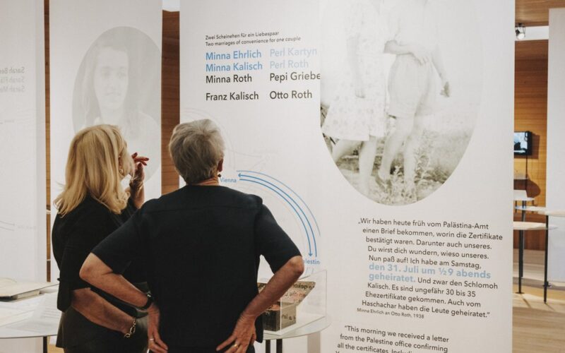 Zwei Frauen stehen vor einer Ausstellungstafel mit historischem Text, Namen und einem Schwarz-Weiß-Foto eines Paares.