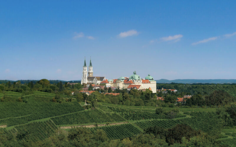 Luftaufnahme eines historischen Klosterkomplexes mit Zwillingstürmen, grünen Kuppeln und umliegenden Weinbergen unter einem klaren blauen Himmel.