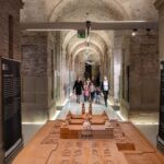 Eine Gruppe von Menschen geht durch einen steinernen Flur mit gewölbten Decken, vorbei an einem komplizierten Holzmodell, das auf einem Tisch in der Mitte steht.