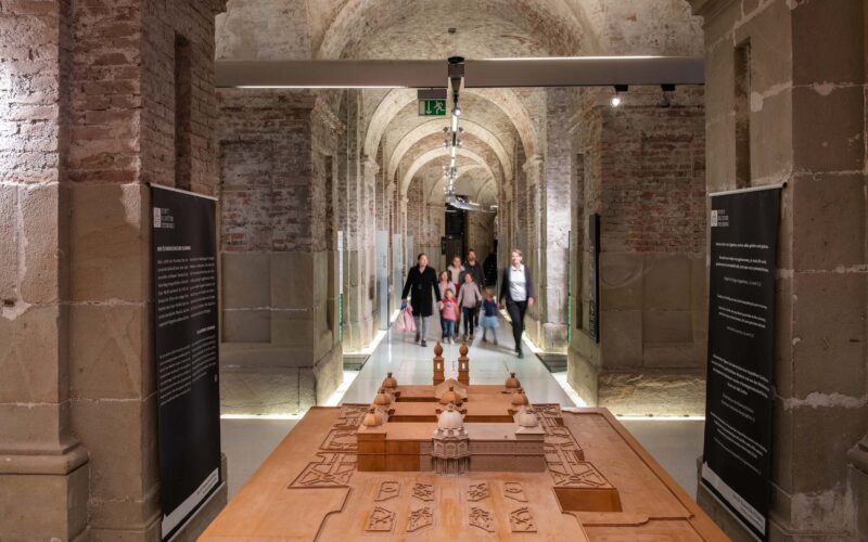 Eine Gruppe von Menschen geht durch einen steinernen Flur mit gewölbten Decken, vorbei an einem komplizierten Holzmodell, das auf einem Tisch in der Mitte steht.