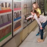 Zwei junge Mädchen interagieren mit einem Ausstellungsstück, indem sie in einem gut beleuchteten Museumsflur eine Schublade herausziehen.