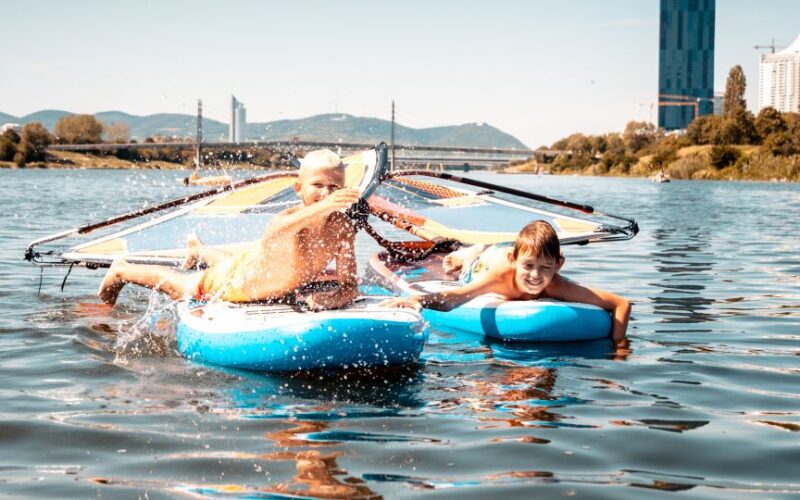 Zwei Kinder spielen auf Paddelbrettern in einem Fluss. Eines hält ein kleines Segel fest. Im Hintergrund sind Gebäude und Hügel unter einem klaren Himmel zu sehen.