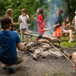 Kinder rösten Essen auf Stöcken über einem Lagerfeuer in einem bewaldeten Außenbereich, während Rauch aufsteigt und andere in der Nähe sitzen oder stehen.