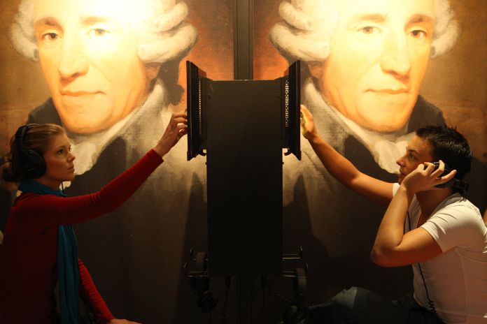 An einer Audiostation sitzen sich zwei Personen mit Kopfhörern gegenüber, im Hintergrund sind große Porträts von Franz Joseph Haydn zu sehen.