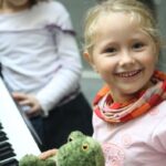 Ein junges Mädchen lächelt in die Kamera, während es einen grünen, ausgestopften Frosch in der Hand hält, während ein anderes Kind im Hintergrund neben einer Klaviertastatur steht.