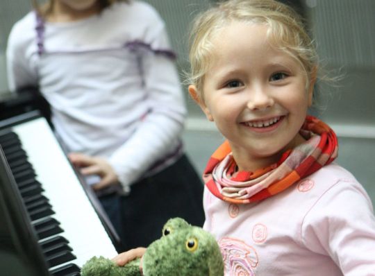 Ein junges Mädchen lächelt in die Kamera, während es einen grünen, ausgestopften Frosch in der Hand hält, während ein anderes Kind im Hintergrund neben einer Klaviertastatur steht.
