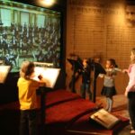 Vier Kinder stehen vor Notenständern und scheinen zu dirigieren, während sie auf einen großen Bildschirm schauen, der ein Orchester in einer museumsähnlichen Umgebung mit Notenblättern an den Wänden zeigt.