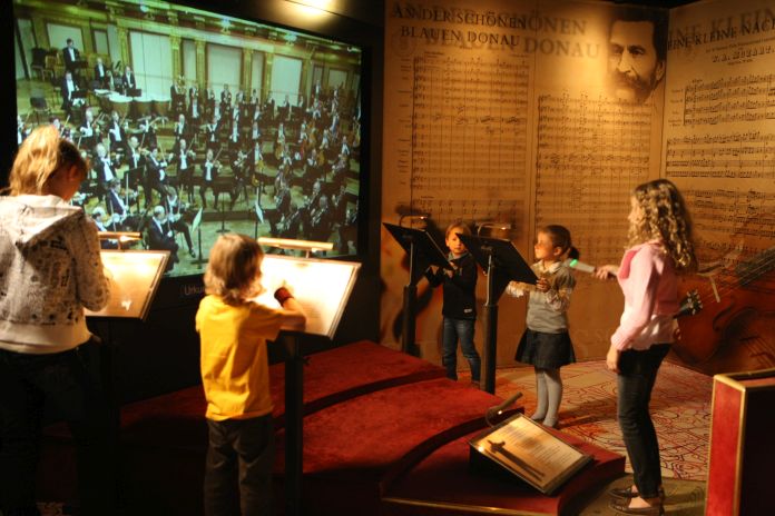 Vier Kinder stehen vor Notenständern und scheinen zu dirigieren, während sie auf einen großen Bildschirm schauen, der ein Orchester in einer museumsähnlichen Umgebung mit Notenblättern an den Wänden zeigt.