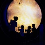 Eine Frau und drei Kinder stehen in Silhouette vor einem großen, beleuchteten, kreisförmigen Display in einem dunklen Raum und berühren dessen Oberfläche.