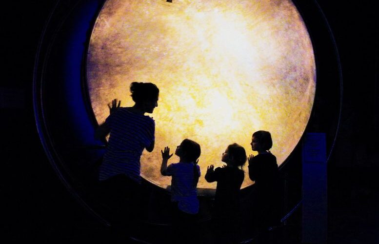 Eine Frau und drei Kinder stehen in Silhouette vor einem großen, beleuchteten, kreisförmigen Display in einem dunklen Raum und berühren dessen Oberfläche.