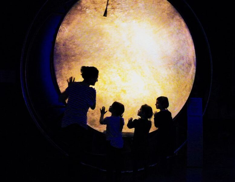 Eine Frau und drei Kinder stehen in Silhouette vor einem großen, beleuchteten, kreisförmigen Display in einem dunklen Raum und berühren dessen Oberfläche.