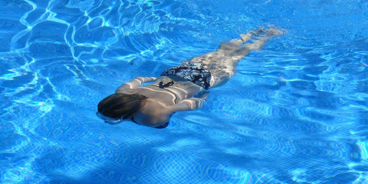Eine Person im Badeanzug schwimmt mit dem Gesicht nach unten in einem klaren blauen Becken, in dem sich das Sonnenlicht auf der Wasseroberfläche spiegelt.