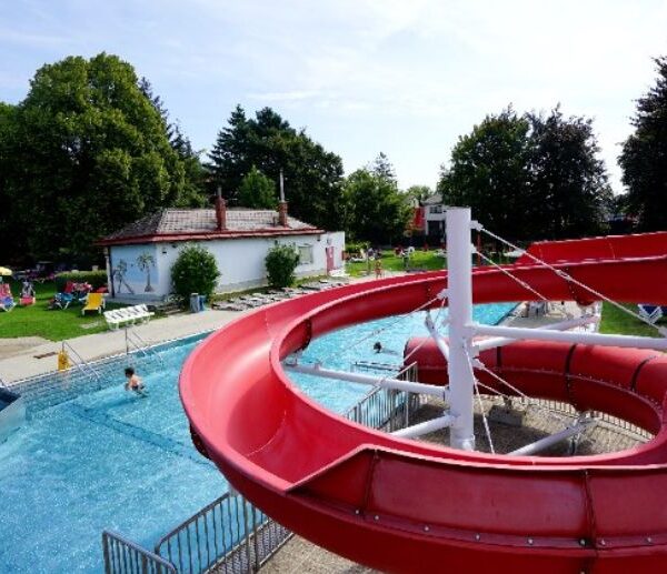 Eine große rote Wasserrutsche führt in einen Swimmingpool im Freien; an sonnigen Tagen entspannt man sich auf dem Rasen und den Liegestühlen unter den Bäumen.
