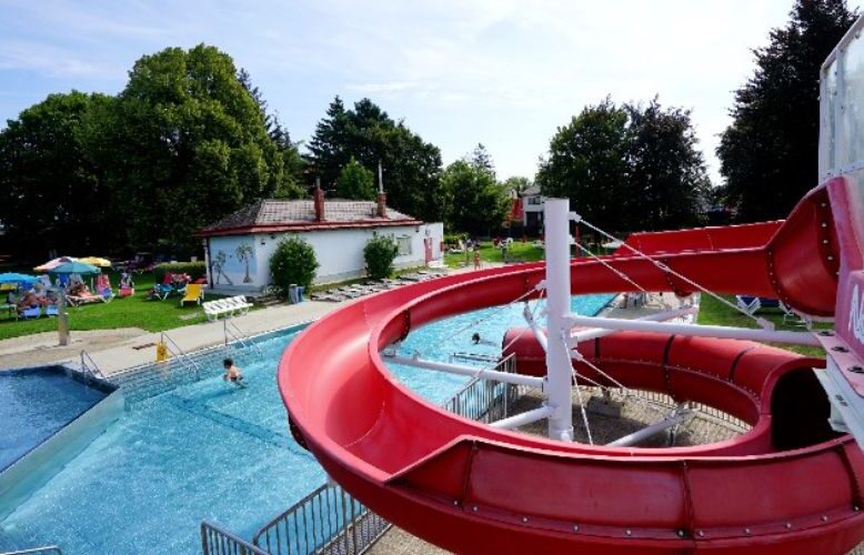 Eine große rote Wasserrutsche führt in einen Swimmingpool im Freien; an sonnigen Tagen entspannt man sich auf dem Rasen und den Liegestühlen unter den Bäumen.
