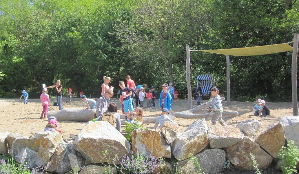 Kinder und Erwachsene versammeln sich und spielen auf einem sandigen, von Felsen gesäumten Spielplatz mit Bäumen und Grünflächen im Hintergrund.