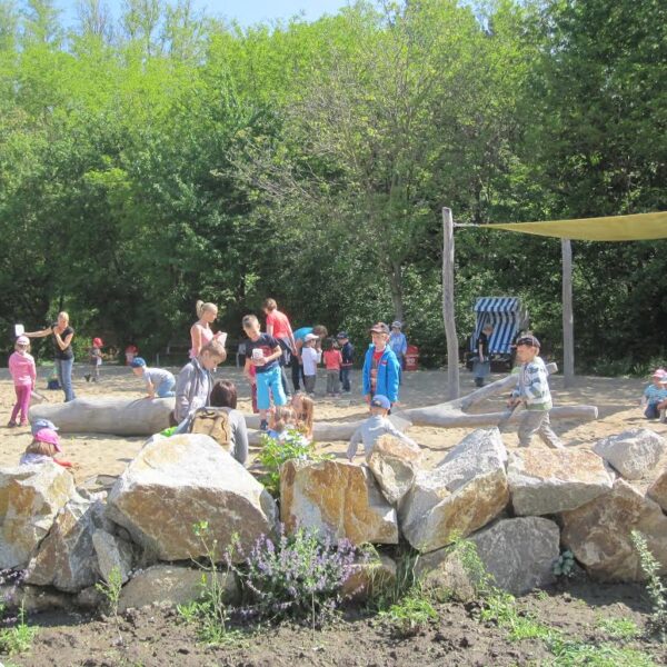 Kinder und Erwachsene versammeln sich und spielen auf einem sandigen, von Felsen gesäumten Spielplatz mit Bäumen und Grünflächen im Hintergrund.