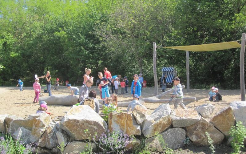 Kinder und Erwachsene versammeln sich und spielen auf einem sandigen, von Felsen gesäumten Spielplatz mit Bäumen und Grünflächen im Hintergrund.