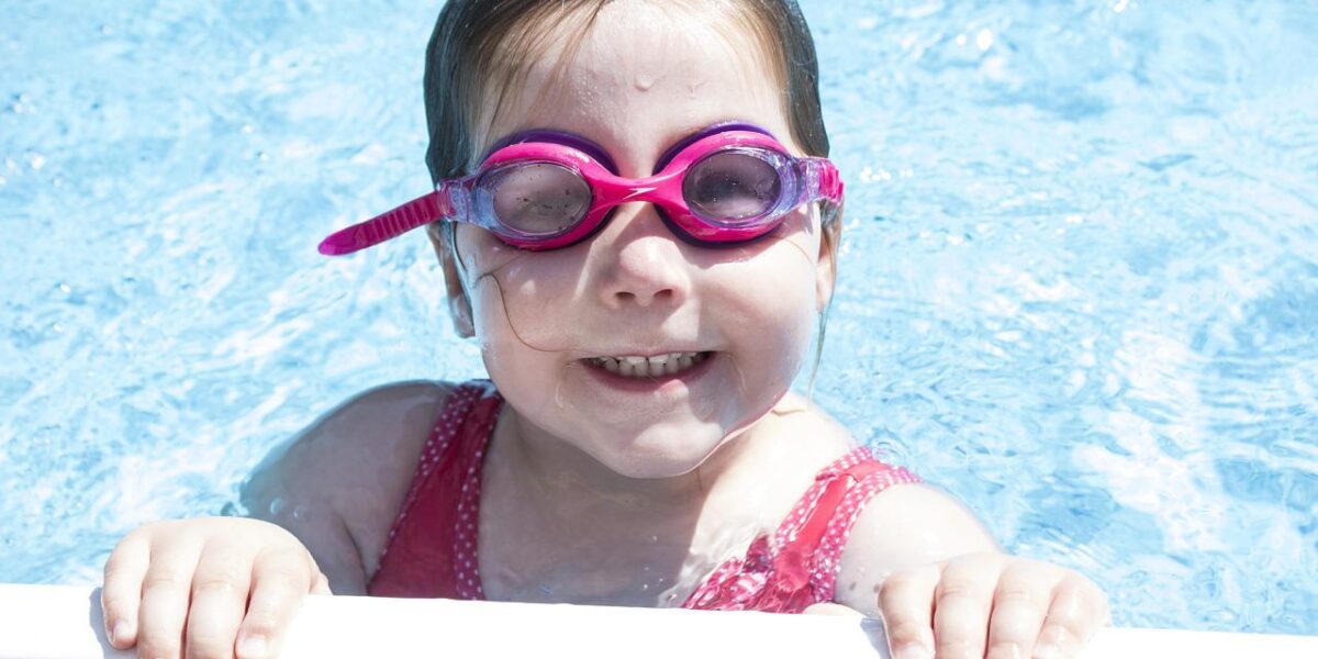 Ein kleines Kind mit rosa Schwimmbrille und rotem Badeanzug lächelt, während es sich am Rand eines Schwimmbeckens festhält, das teilweise in klares blaues Wasser getaucht ist.