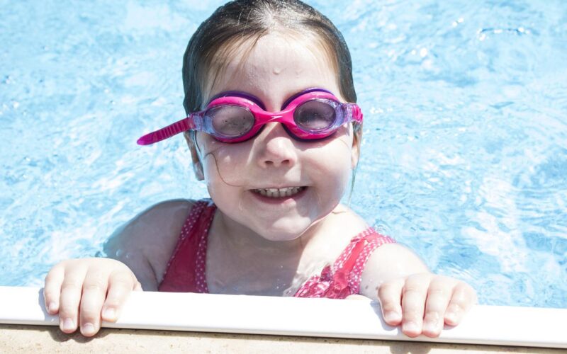Ein kleines Kind mit rosa Schwimmbrille und rotem Badeanzug lächelt, während es sich am Rand eines Schwimmbeckens festhält, das teilweise in klares blaues Wasser getaucht ist.
