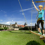 Eine Frau mit Helm und Klettergurt überquert eine Seilbrücke in einem Outdoor-Abenteuerpark, mit einem Schloss und gepflegten Rasenflächen im Hintergrund unter blauem Himmel.