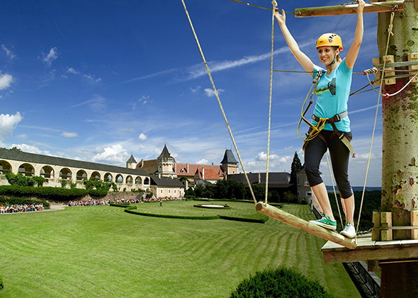Eine Frau mit Helm und Klettergurt überquert eine Seilbrücke in einem Outdoor-Abenteuerpark, mit einem Schloss und gepflegten Rasenflächen im Hintergrund unter blauem Himmel.