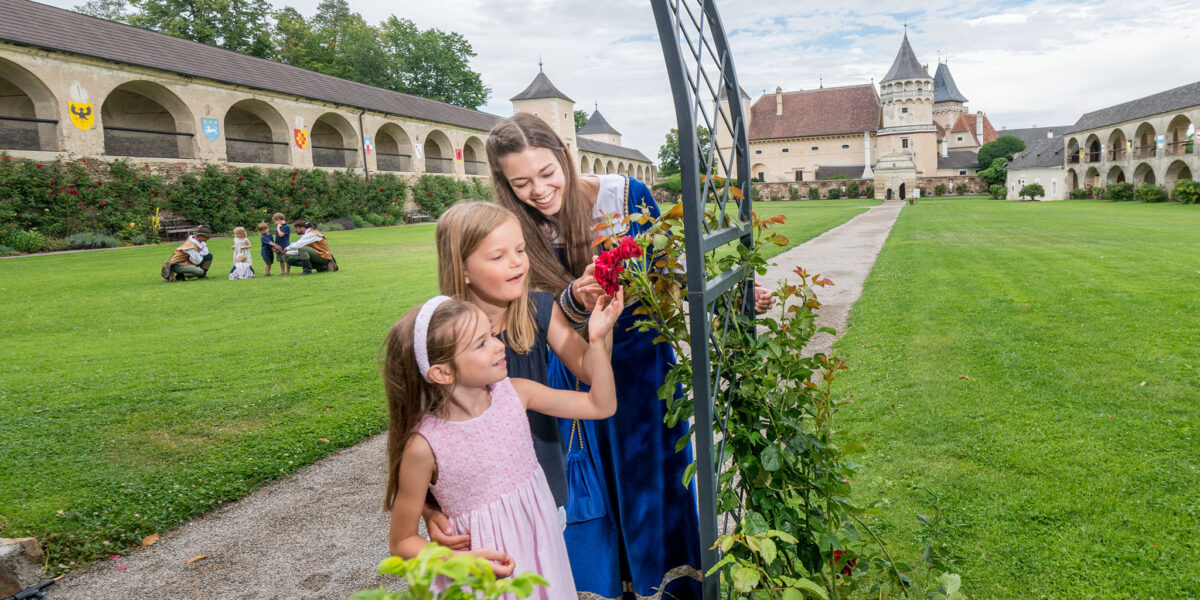 Drei Mädchen in Kleidern stehen an einem Rosenspalier in einem gepflegten Innenhof mit schlossähnlichen Gebäuden im Hintergrund an einem teilweise bewölkten Tag.