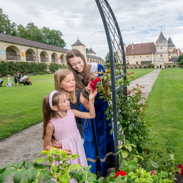 Drei Mädchen in Kleidern stehen an einem Rosenspalier in einem gepflegten Innenhof mit schlossähnlichen Gebäuden im Hintergrund an einem teilweise bewölkten Tag.