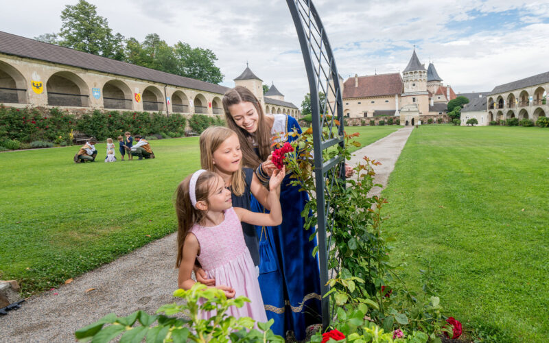 Drei Mädchen in Kleidern stehen an einem Rosenspalier in einem gepflegten Innenhof mit schlossähnlichen Gebäuden im Hintergrund an einem teilweise bewölkten Tag.