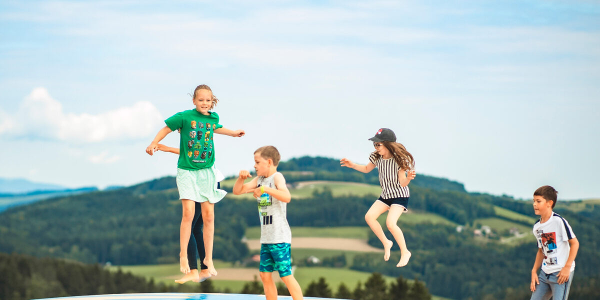 Vier Kinder springen und spielen auf einer aufblasbaren Hüpfburg im Freien, mit grünen Hügeln und einem blauen Himmel im Hintergrund.