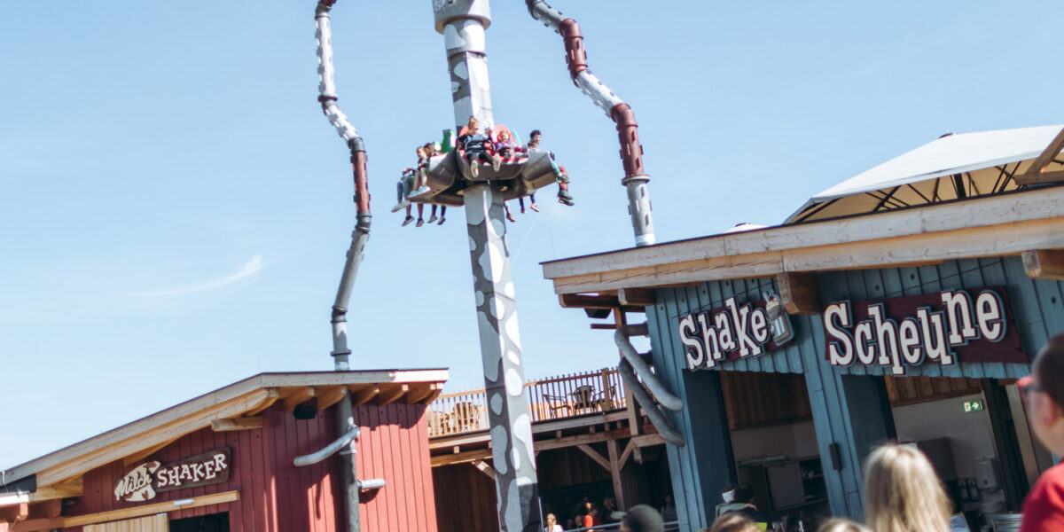 Menschen fahren in einem vertikalen Vergnügungspark, der sie in die Luft hebt und dreht, während in der Nähe Schaulustige und Gebäude mit den Aufschriften "Shake" und "Scheune" zu sehen sind.