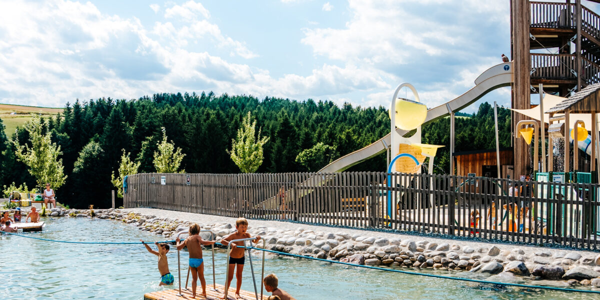 Kinder in Badeanzügen stehen auf einem Holzfloß in einem flachen Freibad in einem Wasserpark, mit einer Rutsche, Wasserspielen und Bäumen im Hintergrund unter einem teilweise bewölkten Himmel.