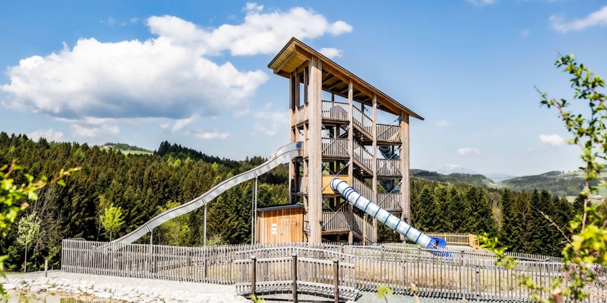 In einem eingezäunten Außenbereich, umgeben von Bäumen und Hügeln unter blauem Himmel, befindet sich ein hohes Holzspielgerät mit mehreren Ebenen und zwei geschlossenen Rutschen.