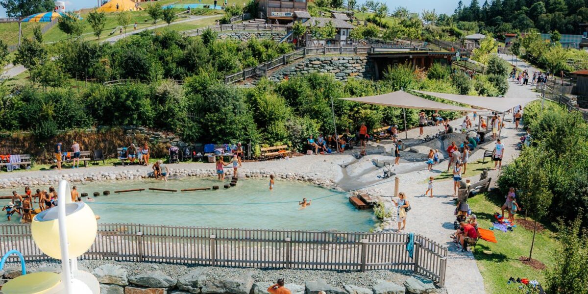 Die Menschen versammeln sich um ein flaches, eingezäuntes Freibad in einem Landschaftspark mit grasbewachsenen Hügeln, Bäumen, Wegen und verschiedenen Parkstrukturen unter einem klaren blauen Himmel.