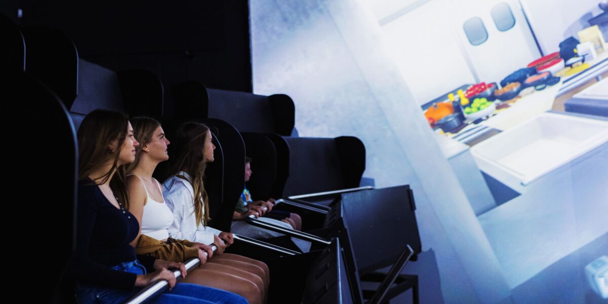 Vier junge Frauen sitzen in einem Fahrsimulator und beobachten einen großen Bildschirm, der eine Innenraumszene mit Küchengeräten zeigt.