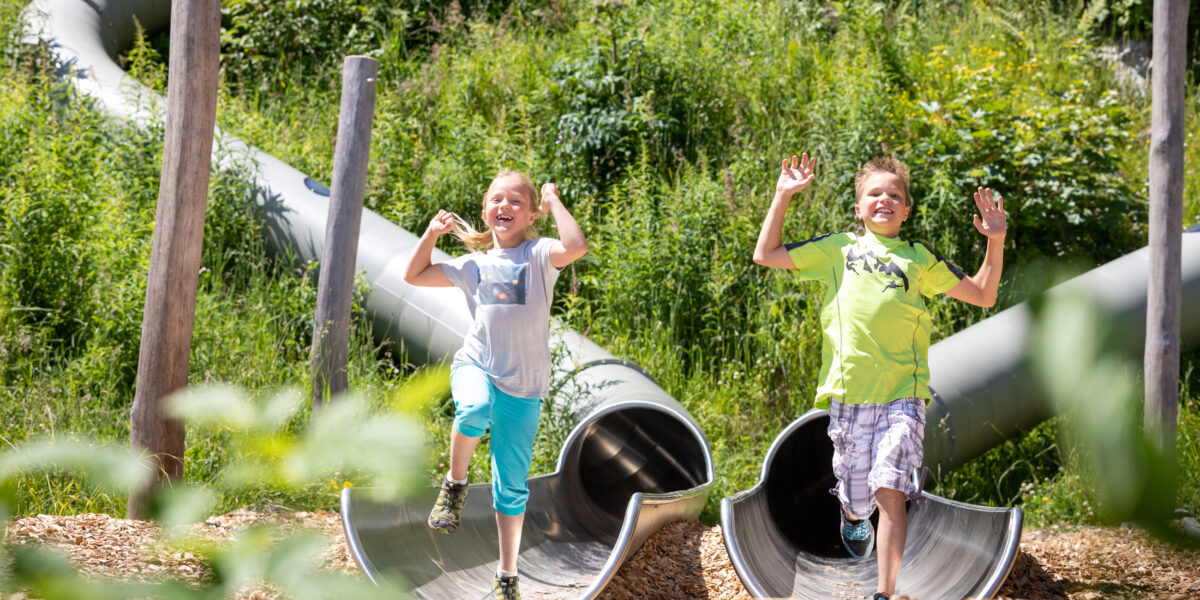 Zwei Kinder, die lächelnd die Arme heben, als sie die Metallröhrenrutsche auf einem Spielplatz im Freien verlassen, der von viel Grün umgeben ist.