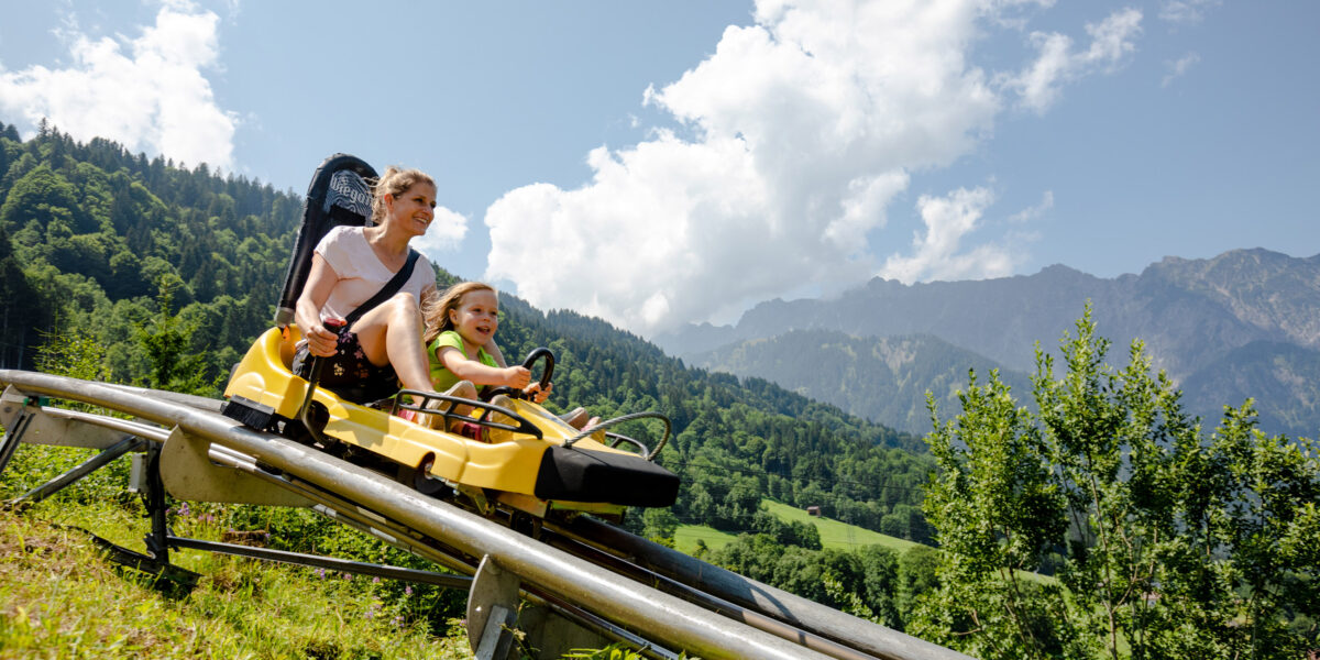 Ein Erwachsener und ein Kind fahren auf einer gelben Alpenachterbahn einen Hügel hinunter, mit grünen Bäumen und Bergen im Hintergrund unter einem teilweise bewölkten Himmel.