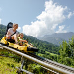 Ein Erwachsener und ein Kind fahren auf einer gelben Alpenachterbahn einen Hügel hinunter, mit grünen Bäumen und Bergen im Hintergrund unter einem teilweise bewölkten Himmel.