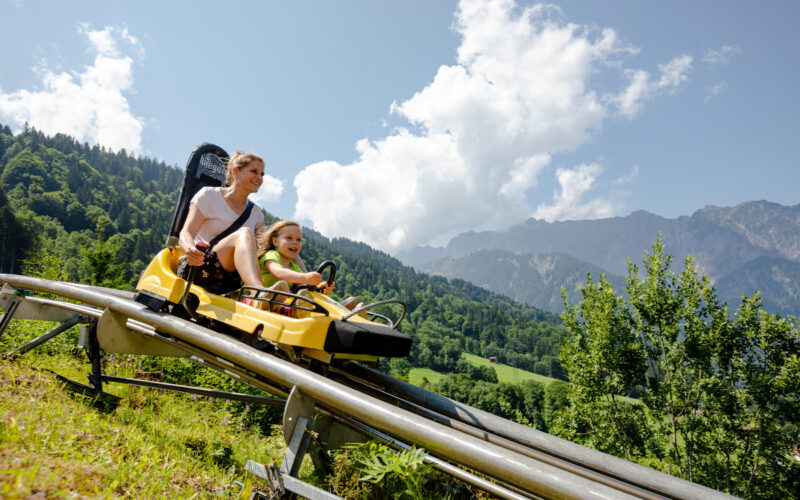 Ein Erwachsener und ein Kind fahren auf einer gelben Alpenachterbahn einen Hügel hinunter, mit grünen Bäumen und Bergen im Hintergrund unter einem teilweise bewölkten Himmel.