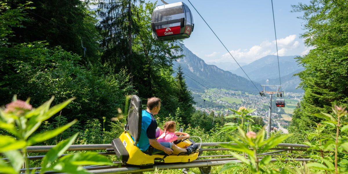 Ein Mann und ein Kind fahren mit einem gelben Alpine Coaster einen grünen Hügel hinunter, während über ihnen Seilbahnen fahren und im Hintergrund ein Bergtal zu sehen ist.