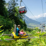 Ein Mann und ein Kind fahren mit einem gelben Alpine Coaster einen grünen Hügel hinunter, während über ihnen Seilbahnen fahren und im Hintergrund ein Bergtal zu sehen ist.