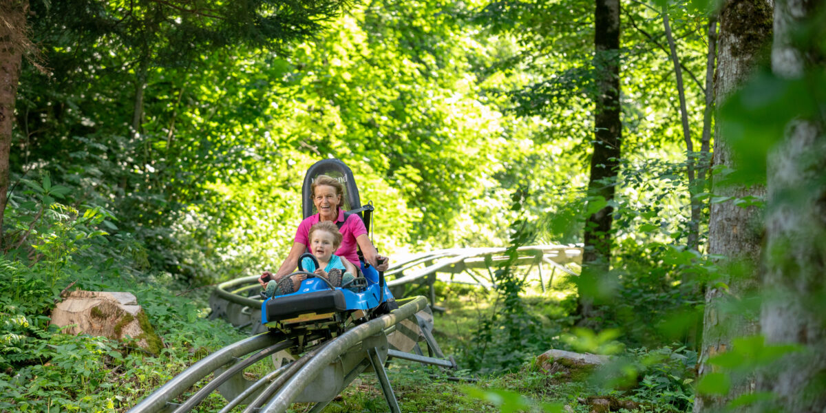 Ein Erwachsener und ein Kind fahren an einem sonnigen Tag mit einer Achterbahn durch einen üppigen grünen Wald.