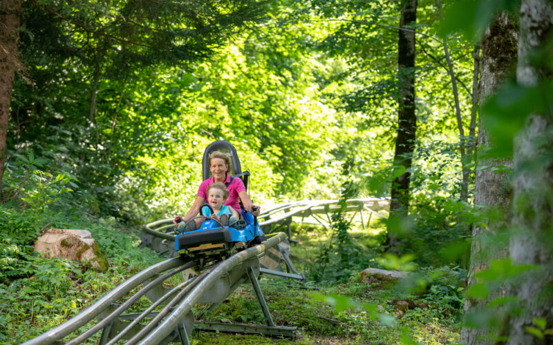 Ein Erwachsener und ein Kind fahren an einem sonnigen Tag mit einer Achterbahn durch einen üppigen grünen Wald.