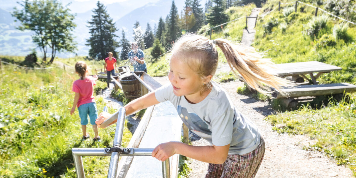Ein junges Mädchen dreht an einem sonnigen Tag im Freien eine Wasserpumpe mit Metallspirale, während im grasbewachsenen, bergigen Hintergrund Kinder spielen.